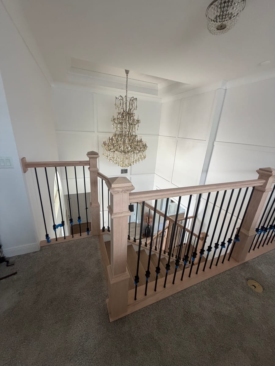 White oak staircase with black iron balusters beneath a crystal chandelier, upstairs landing view
